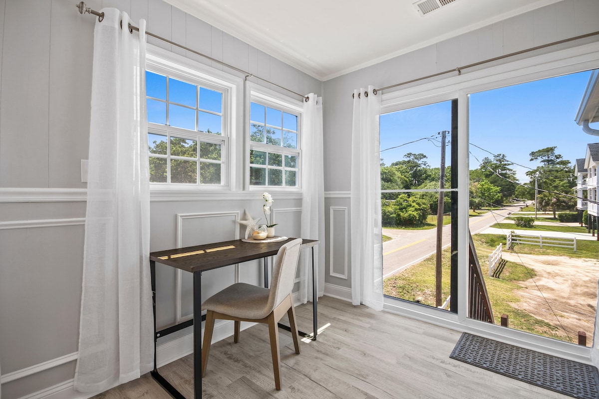 A bright corner workspace features a simple desk with a chair and is illuminated by natural light from large windows. Sheer white curtains frame the windows, providing a soft contrast to the light grey walls. A view of the surrounding greenery and street is visible.