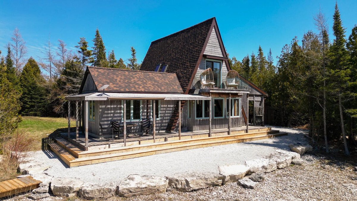 A renovated A-frame retreat is showcased under clear blue skies. The property features a wooden deck with outdoor seating and a well manicured gravel area, surrounded by trees. The charming exterior highlights a mix of wooden and stone elements, inviting guests to enjoy the serene surroundings.