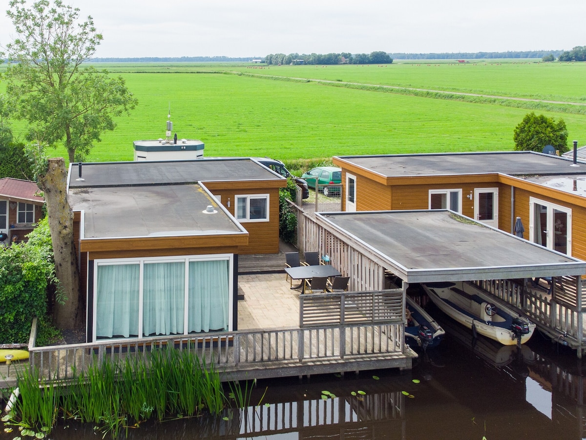 The chalet is viewed from above, showcasing a wooden exterior and large windows, situated alongside a tranquil waterway. A spacious terrace is present, accompanied by a covered boathouse housing a small boat. Expansive green fields are visible in the background.