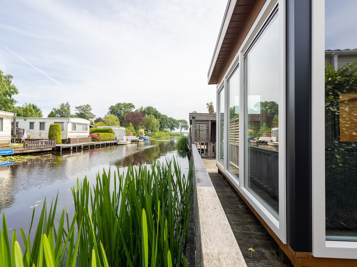 The image captures a serene view along a wooden deck lined by tall green reeds, leading to a tranquil waterway. On the opposite bank, charming chalets are visible among lush greenery, enhancing the peaceful setting. The sky above is bright with soft clouds.