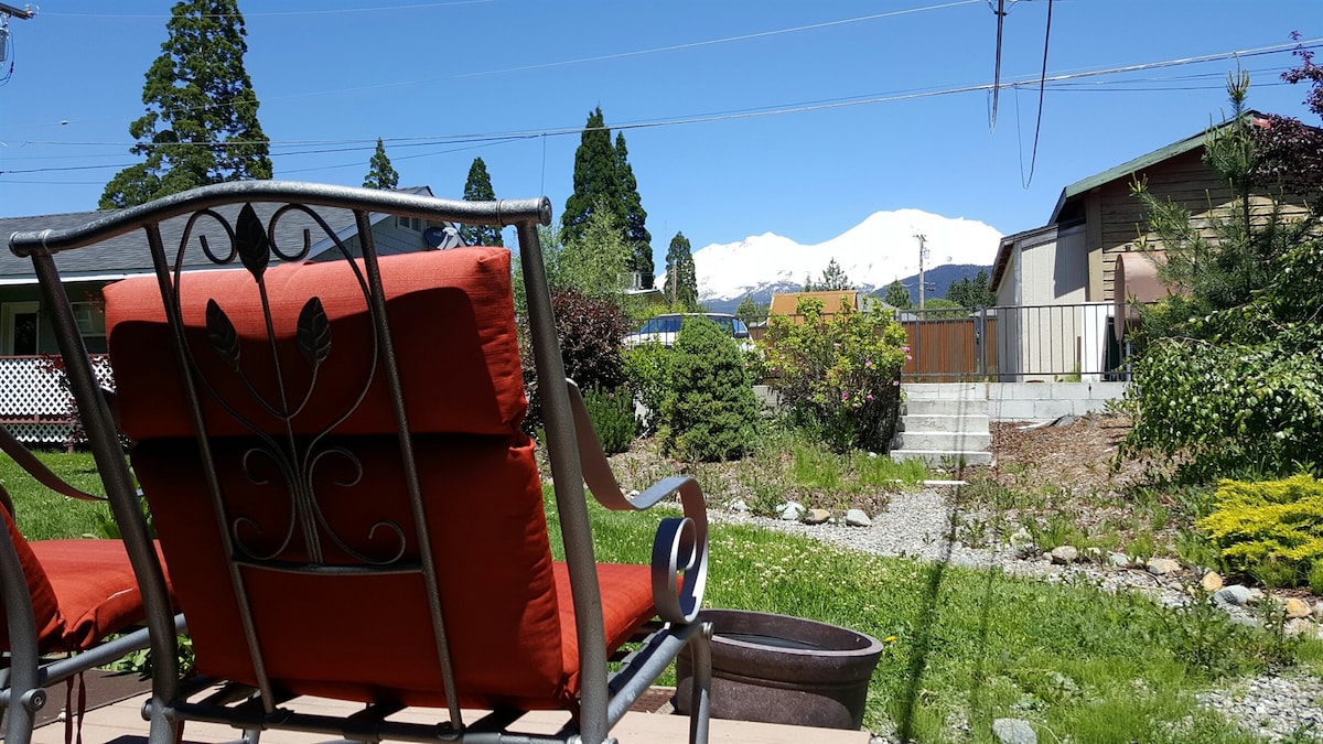 A shaded outdoor seating area features a metal chair with bright orange cushions, facing a well-maintained garden. In the background, snow-capped mountains are visible under a clear blue sky, while lush greenery surrounds the space.