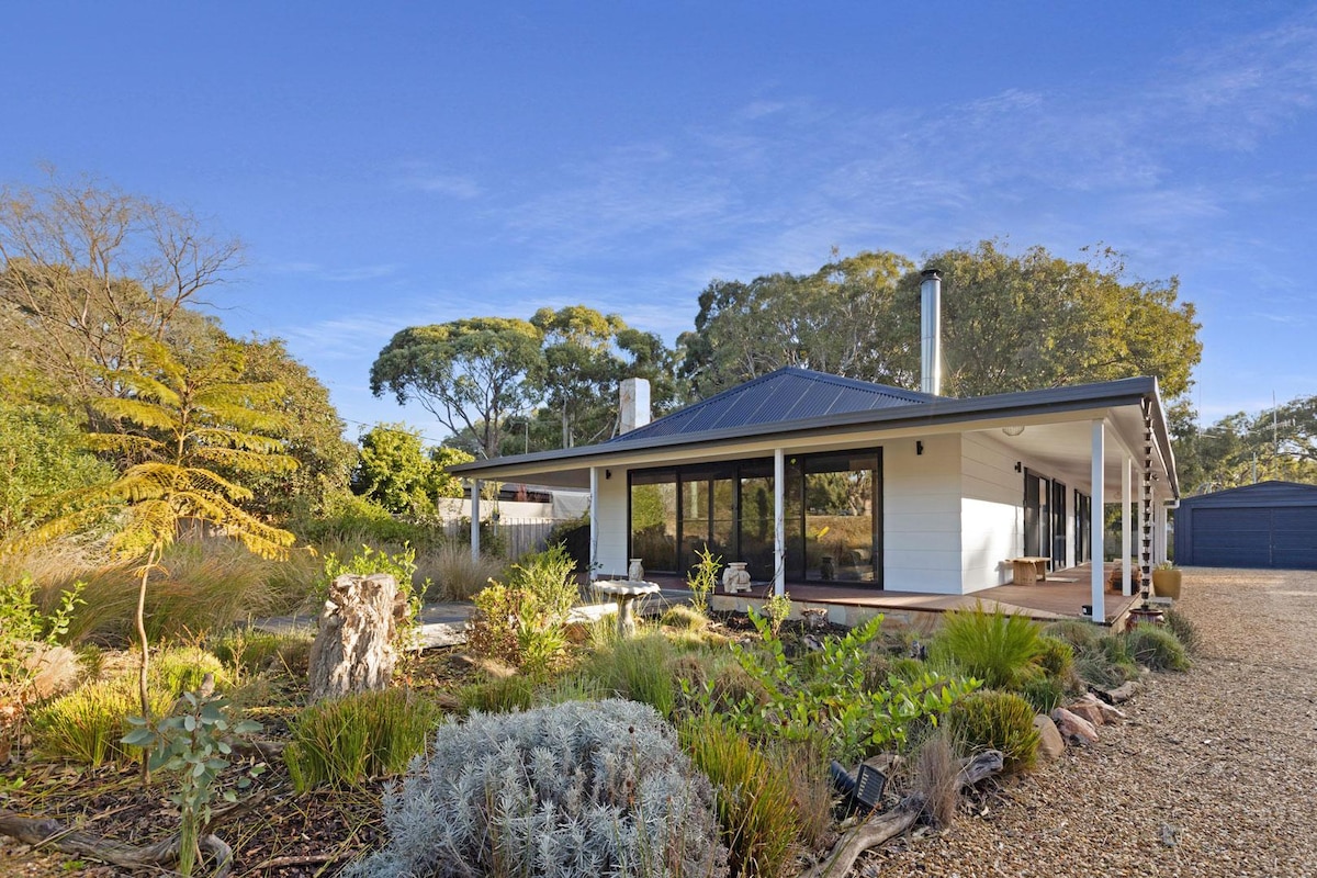A modern single-story house is surrounded by a natural landscape, featuring a variety of plants and trees. Large glass doors create a connection to the outdoors, while a gravel pathway leads to the entrance. The roof is finished in dark metal, enhancing the architectural appeal.