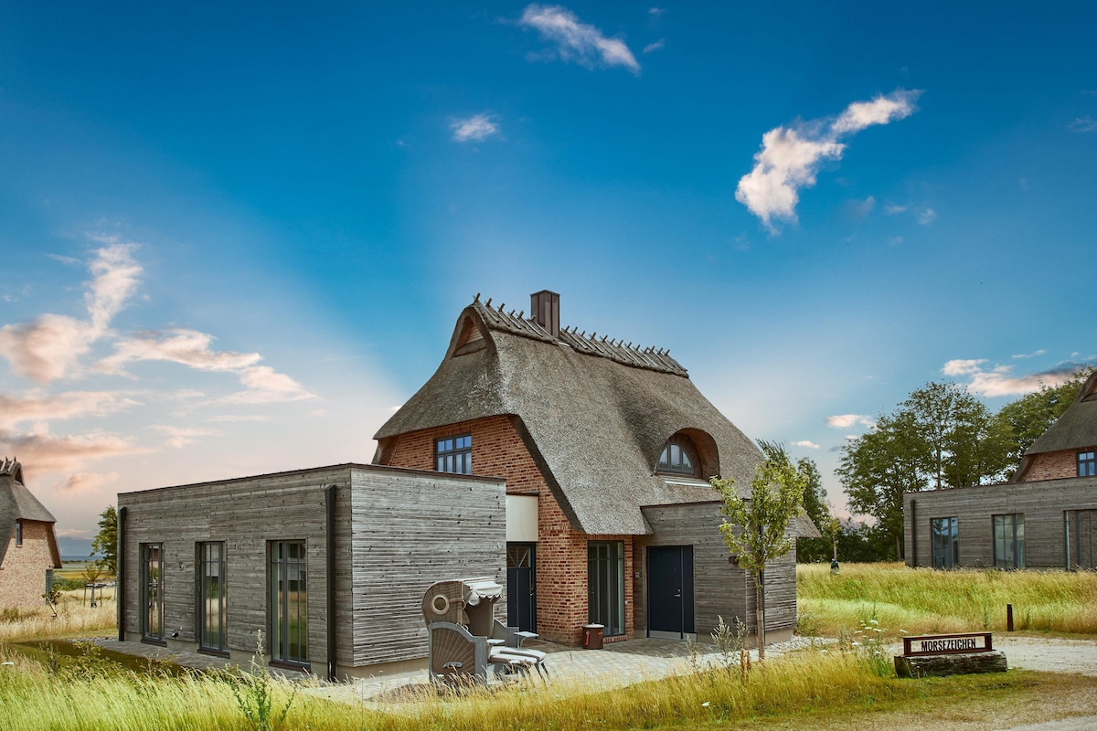 A spacious studio house features a thatched roof and a combination of wood and brick exterior. The structure is surrounded by open grassland, with a clear sky above. A barbecue is positioned beside the entrance, and sun loungers are visible on the terrace.