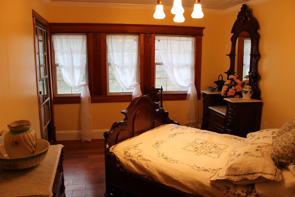 A cozy bedroom features a high-backed wooden bed draped with a patterned coverlet. Soft morning light filters through white sheer curtains adorning two windows. A vintage dresser and mirror, along with a side table and china bowl, complete the room's inviting atmosphere.