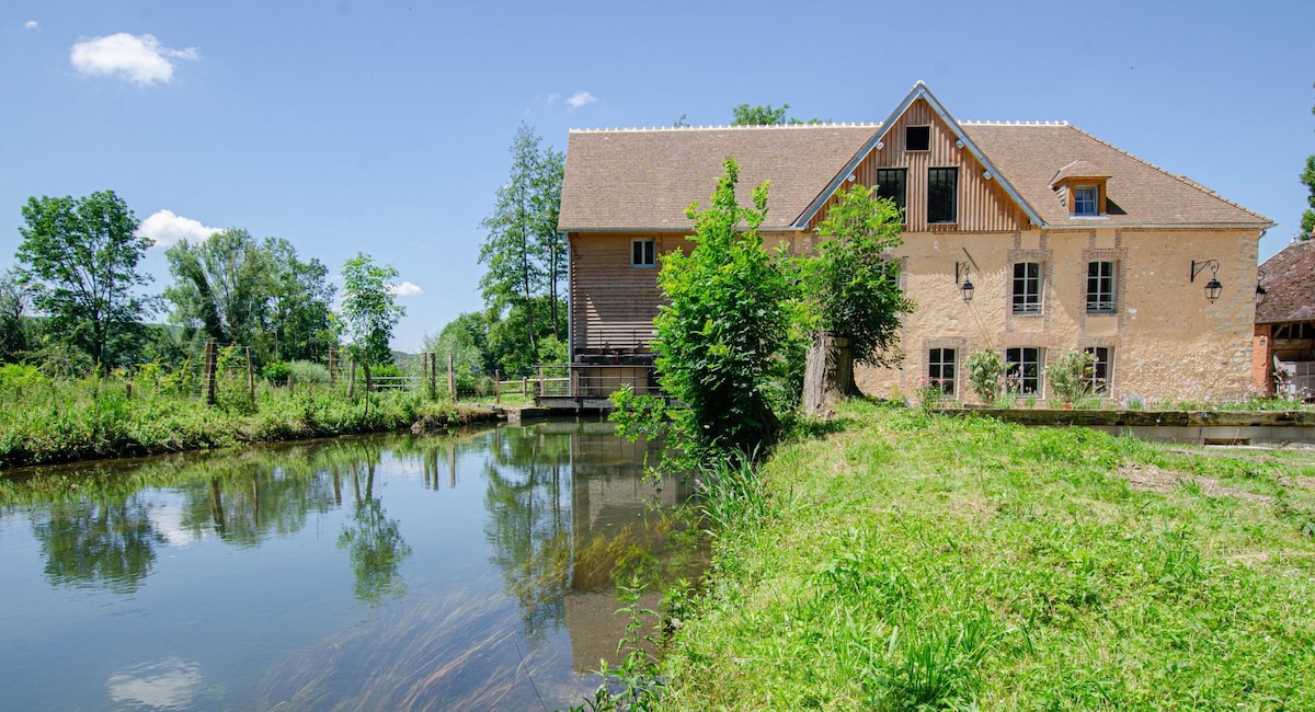 A traditional mill building with wooden and stone features is set beside a calm water body, surrounded by lush greenery. Large windows offer views of the serene landscape, enhancing the peaceful environment. The property showcases a blend of historical charm and natural beauty.