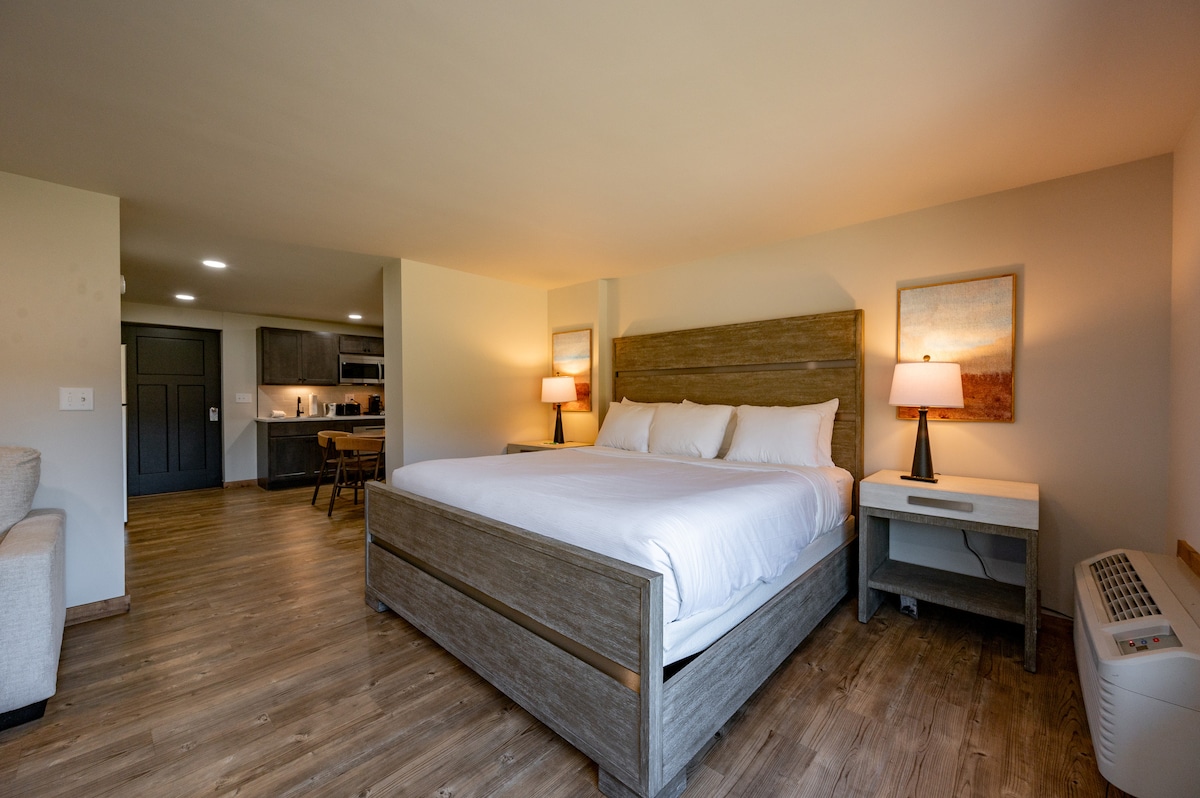A spacious sleeping area features a plush King bed with crisp linens. The room is illuminated by two bedside lamps, casting a soft glow. A neutral color palette complements the wooden furniture. The open layout provides a view of the adjoining kitchen and dining area.