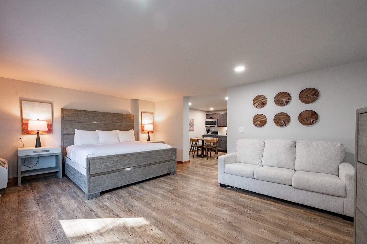 A spacious room features a King bed with light-colored linens and two bedside lamps, providing soft illumination. A plush Queen sleeper sofa is positioned nearby, with a wooden dining table visible in the background. Wood-like flooring contributes to the serene environment.