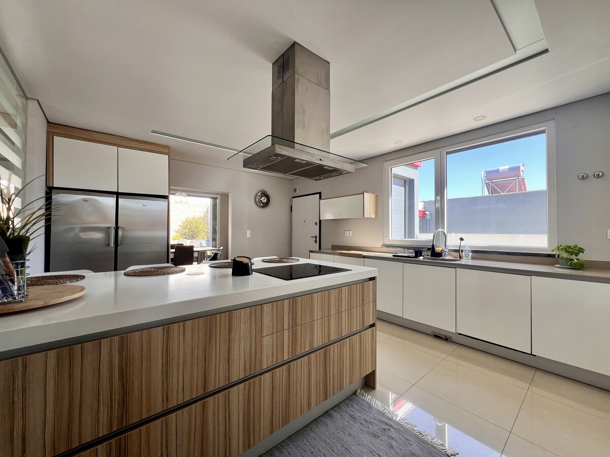 A modern kitchen features sleek cabinetry with wooden accents and a spacious island countertop. Stainless steel appliances are positioned for convenience, while natural light flows through large windows, brightening the open layout.
