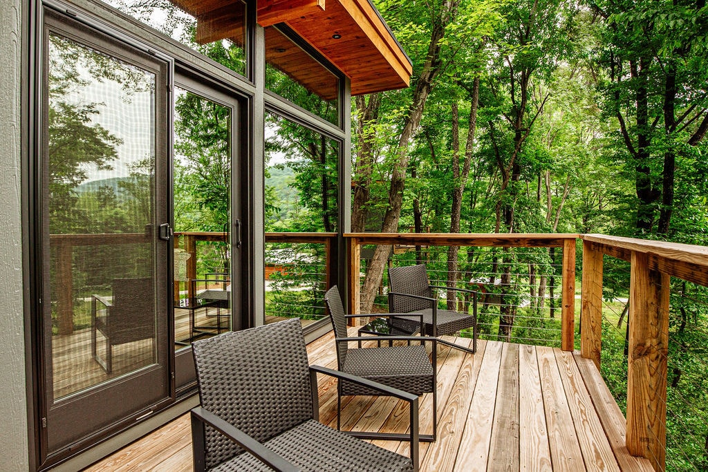 A private deck features wooden flooring and railings, surrounded by a lush landscape of trees. Four wicker lounge chairs are arranged to provide comfortable seating while enjoying the natural environment. Expansive windows visible from the deck allow for ample natural light in the suite.