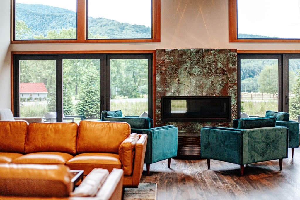 The common lounge area is designed with large windows, offering abundant natural light and sweeping views of the surrounding landscape. A modern gas fireplace is positioned centrally, surrounded by comfortable seating, including a spacious leather sofa and stylish armchairs in various hues.