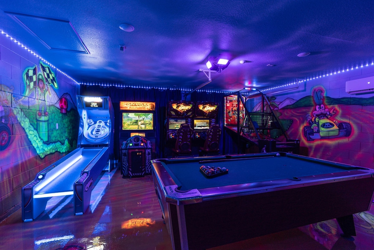 A vibrant game room is illuminated with colorful lighting, featuring a pool table and various arcade machines. Decorative murals adorn the walls, adding a playful ambiance. The space invites guests to enjoy gaming and entertainment in a dynamic setting.