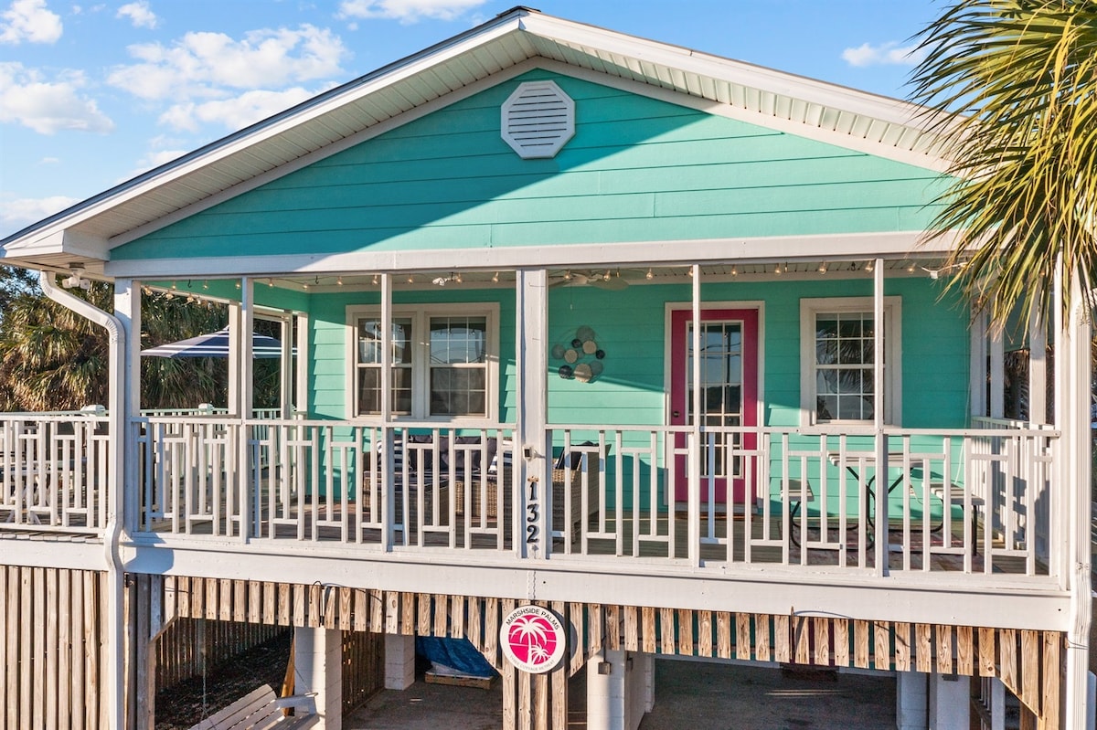 A charming cottage is displayed in vibrant turquoise, featuring a wraparound porch adorned with white railings. The entrance is highlighted by a pink door, surrounded by multiple seating areas. Lush palm trees and blue skies provide a serene backdrop for this inviting retreat.