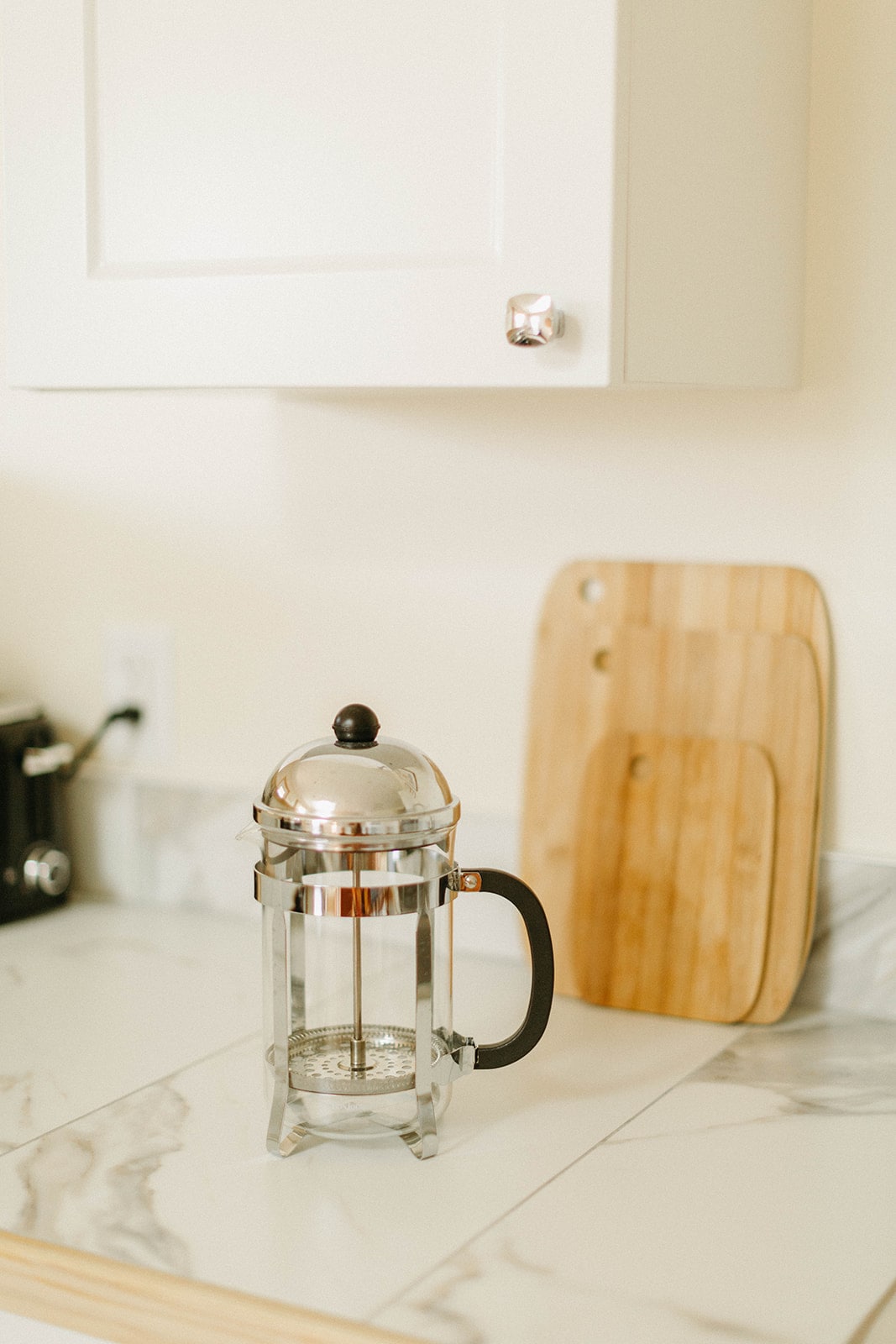 A French press made of glass and metal is positioned on a marble countertop. Behind it, a wooden cutting board leans against a wall-mounted cabinet, adding warmth to the kitchen space. Clean lines and neutral tones create an inviting environment.