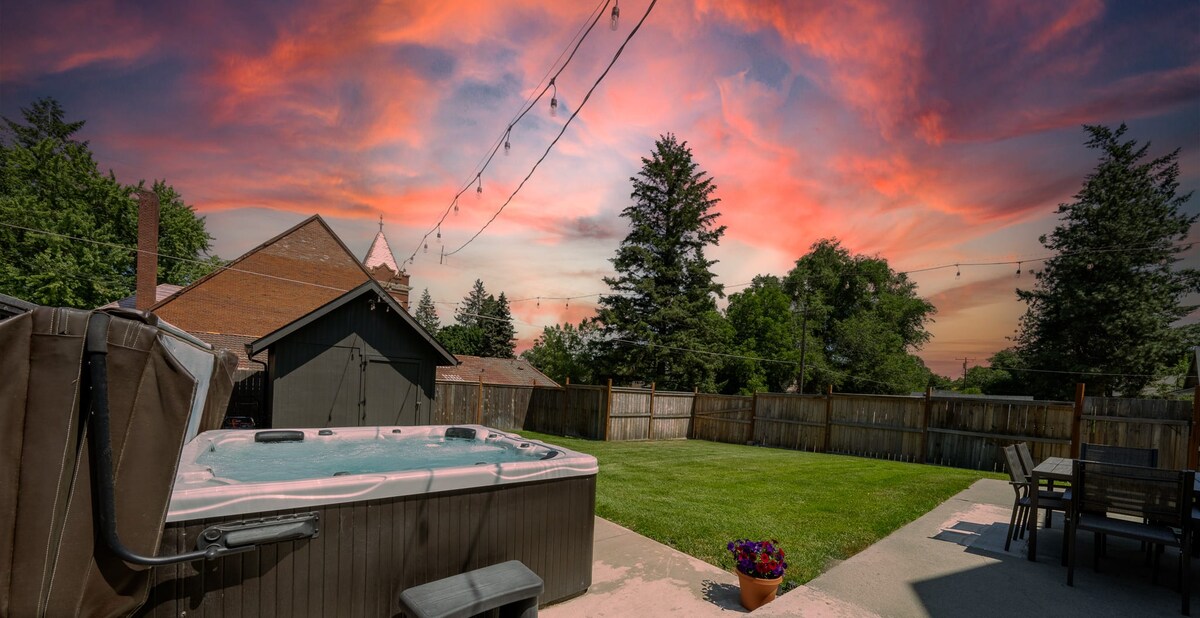 A fenced backyard showcases a hot tub next to a concrete pad. Grassy areas are present, surrounded by trees and a wooden fence. String lights are visible overhead against a colorful sunset sky, enhancing the inviting outdoor space.
