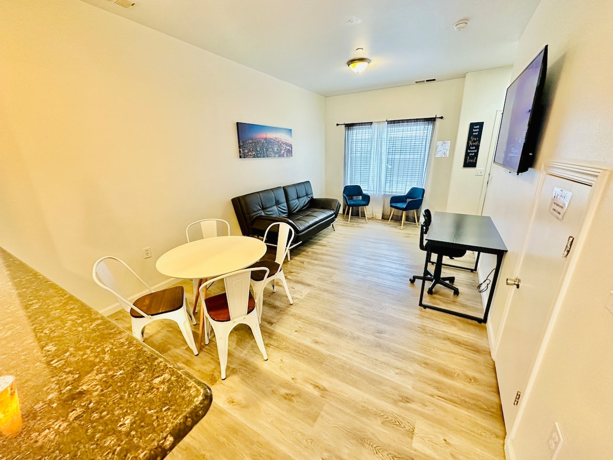 An open-concept living area features a sleek black sofa and two blue chairs positioned near a window. A round dining table with white chairs and a work desk are included. Light wood flooring and a mounted television enhance the modern atmosphere.