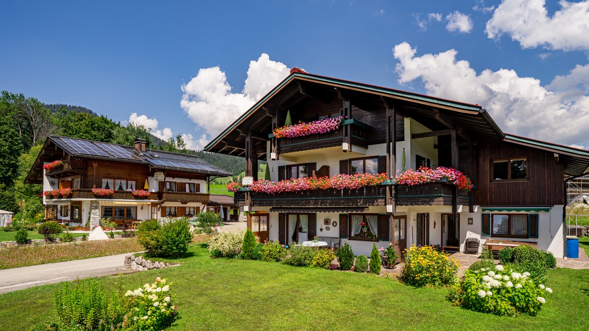 A charming chalet-style building with flower-filled balconies is presented against a clear blue sky. Lush green grass surrounds the structure, complemented by neatly arranged flower beds. Solar panels are visible on the roof, highlighting environmentally friendly features.