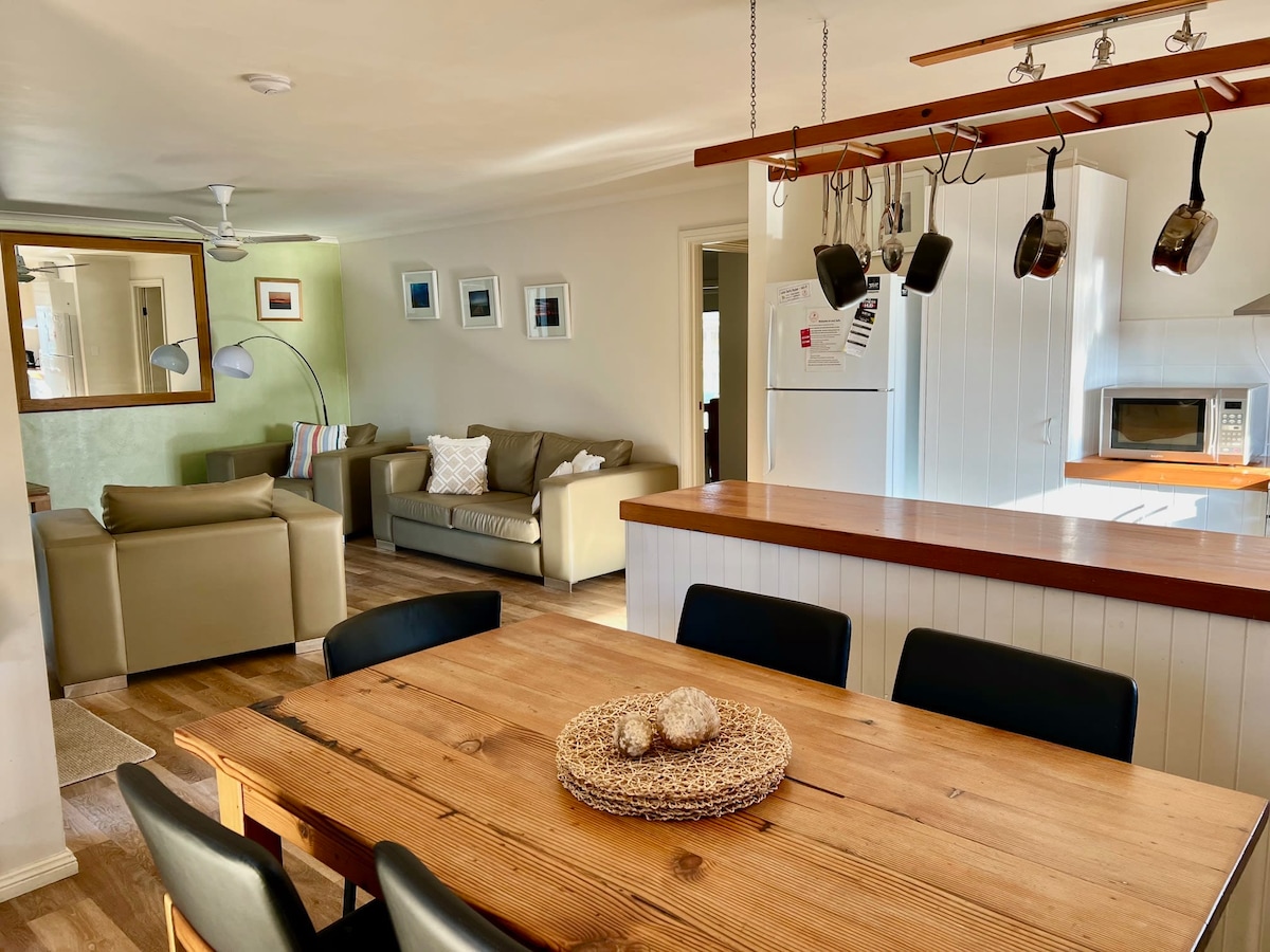 An open-plan living area showcases a wooden dining table in the foreground, surrounded by black chairs. A light-colored sofa and armchair are visible in the background, along with a ceiling fan and ample natural light from nearby windows.
