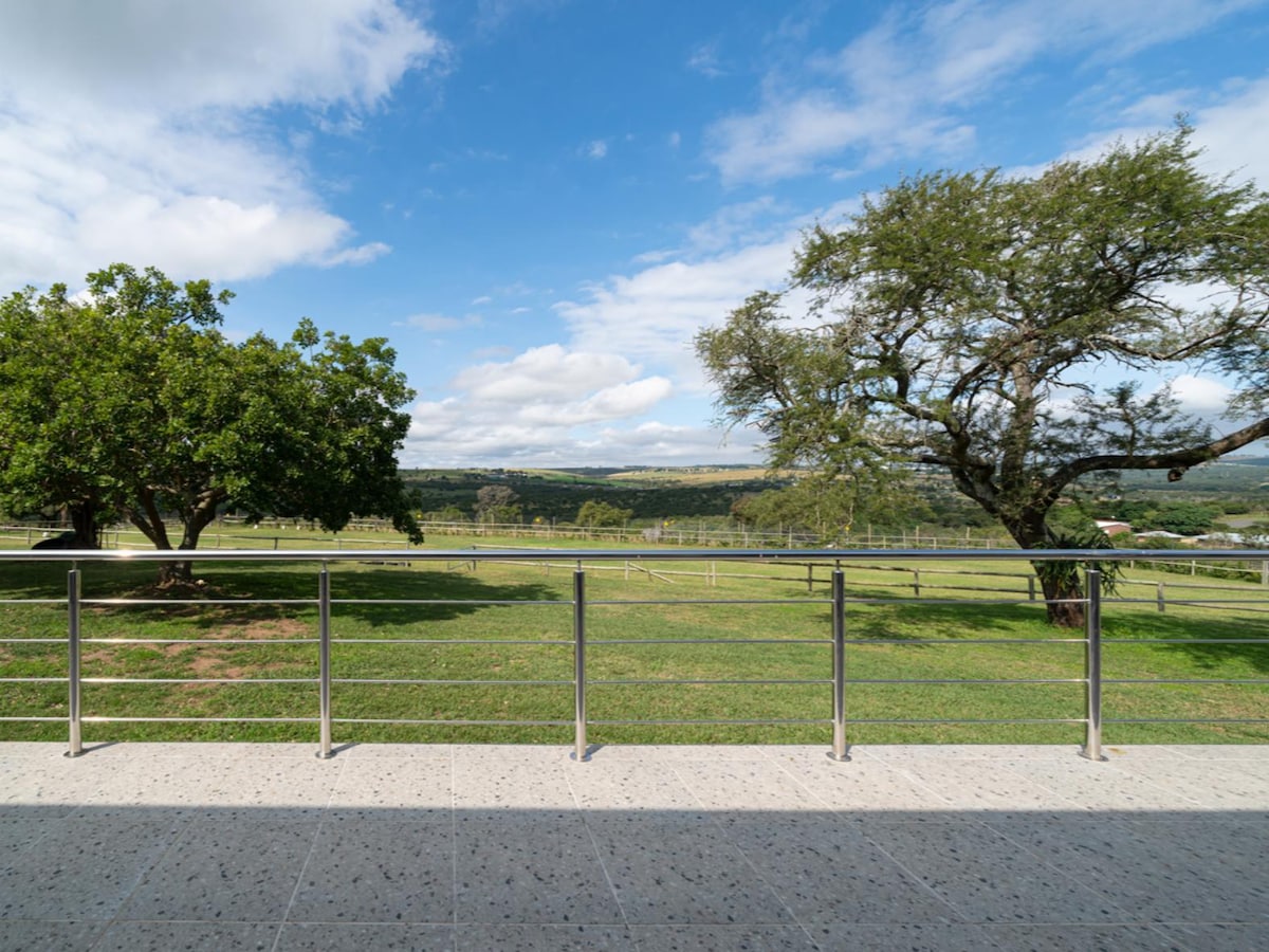 A spacious balcony features a metal railing overlooking a verdant landscape. Lush green fields extend into the distance, framed by scattered trees beneath a partially cloudy sky. The serene environment invites relaxation and offers unobstructed views of the natural surroundings.