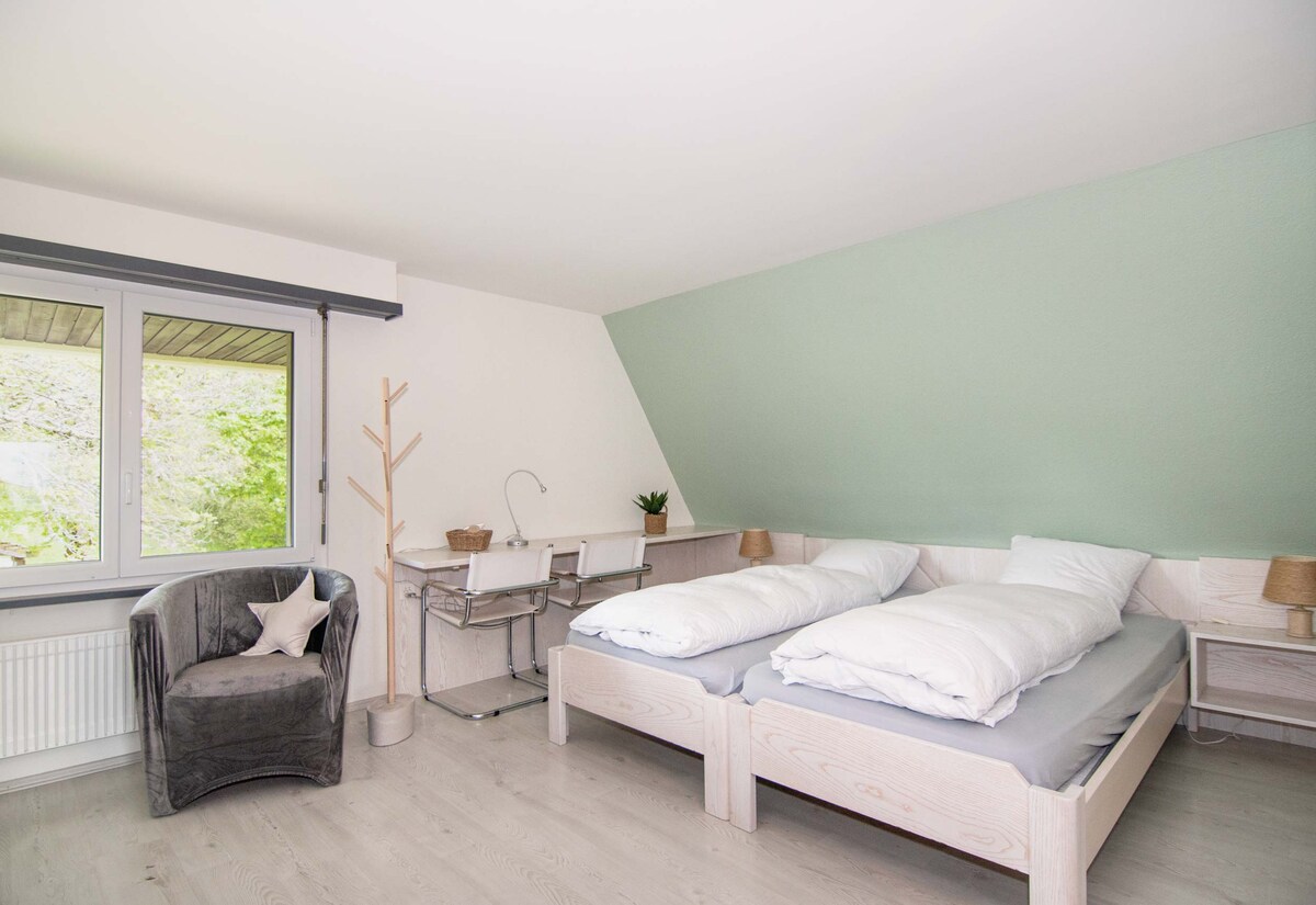 A bright guest room features twin beds with light bedding and a spacious desk beside a large window. A comfortable chair sits in one corner, complemented by neutral decor. Natural light floods the room, accentuating the soothing green wall and airy atmosphere.