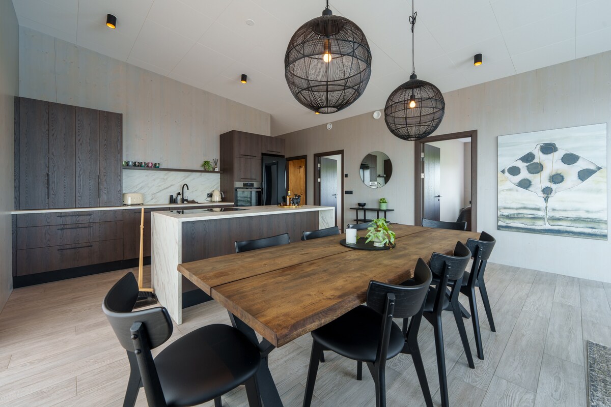 A modern kitchen and dining area features a large wooden table surrounded by black chairs. Suspended wire pendant lights illuminate the space, where sleek cabinetry and a countertop complement the open layout. A piece of artwork adorns one wall, adding a touch of style.