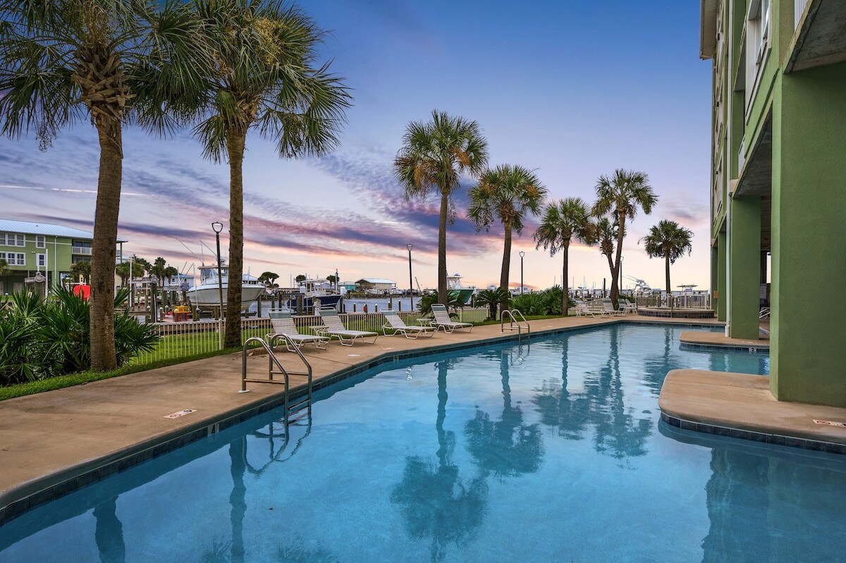 A serene swimming pool is framed by palm trees, offering a clear view of the water. Lounge chairs line the poolside, inviting relaxation. The vibrant sunset sky features soft hues of purple and orange, enhancing the tranquil atmosphere.