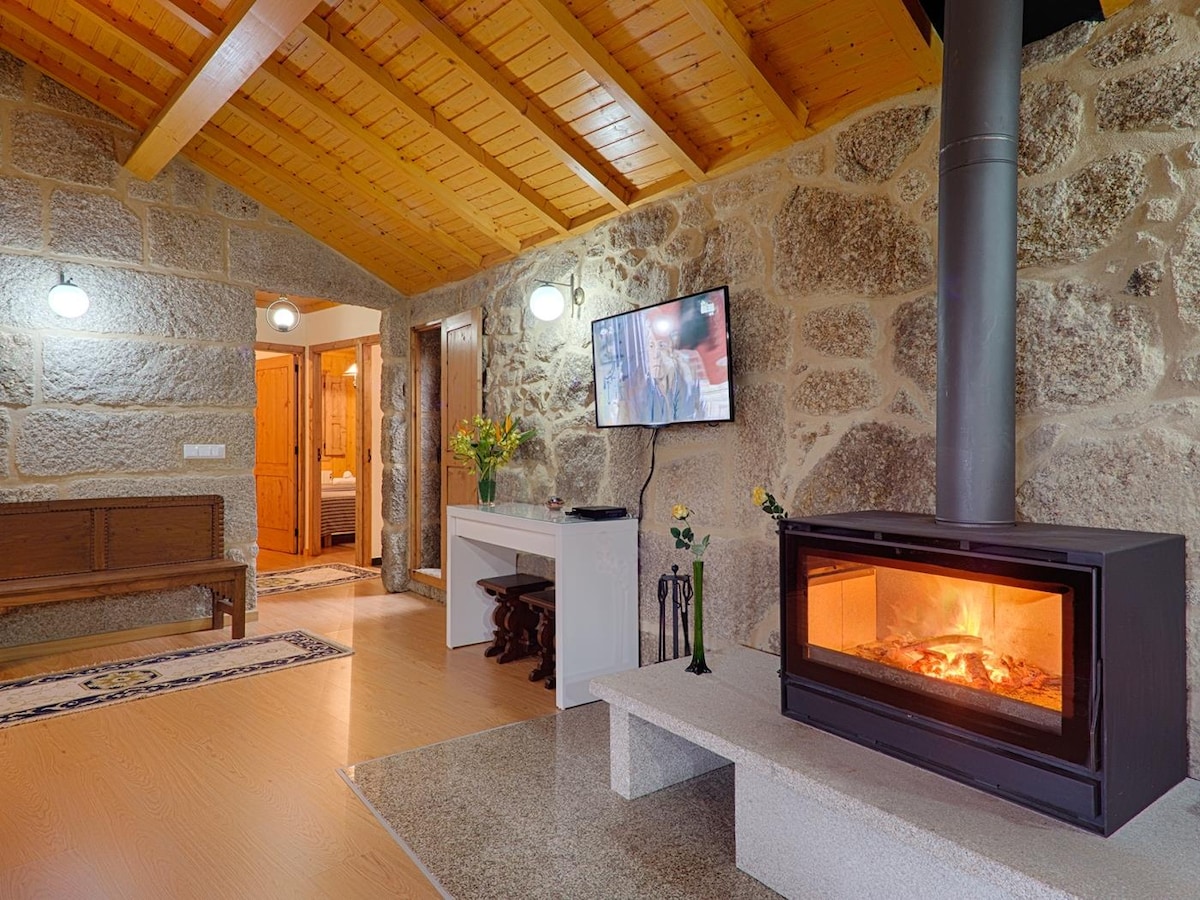 A cozy living area features exposed stone walls and a wooden beam ceiling, creating a warm ambiance. A modern fireplace sits in front of a sleek television, while a console table and decorative flowers enhance the space. The room connects to a doorway leading to additional areas.