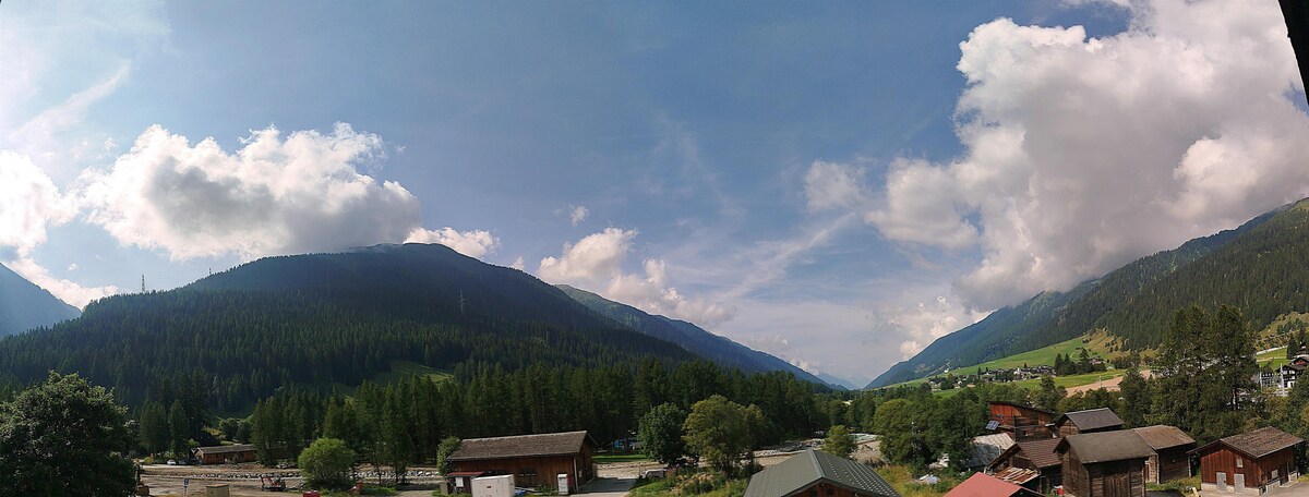 A panoramic view showcases expansive mountain ranges and lush green forests under a partly cloudy sky. The landscape includes scattered wooden buildings nestled among trees, with a wide valley stretching into the distance.
