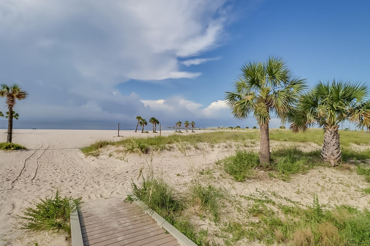 A sandy beach pathway leads through tufts of grass and clusters of palm trees, showcasing the calming blue of the ocean in the distance. The sky is partially cloudy, adding depth to the coastal scenery.