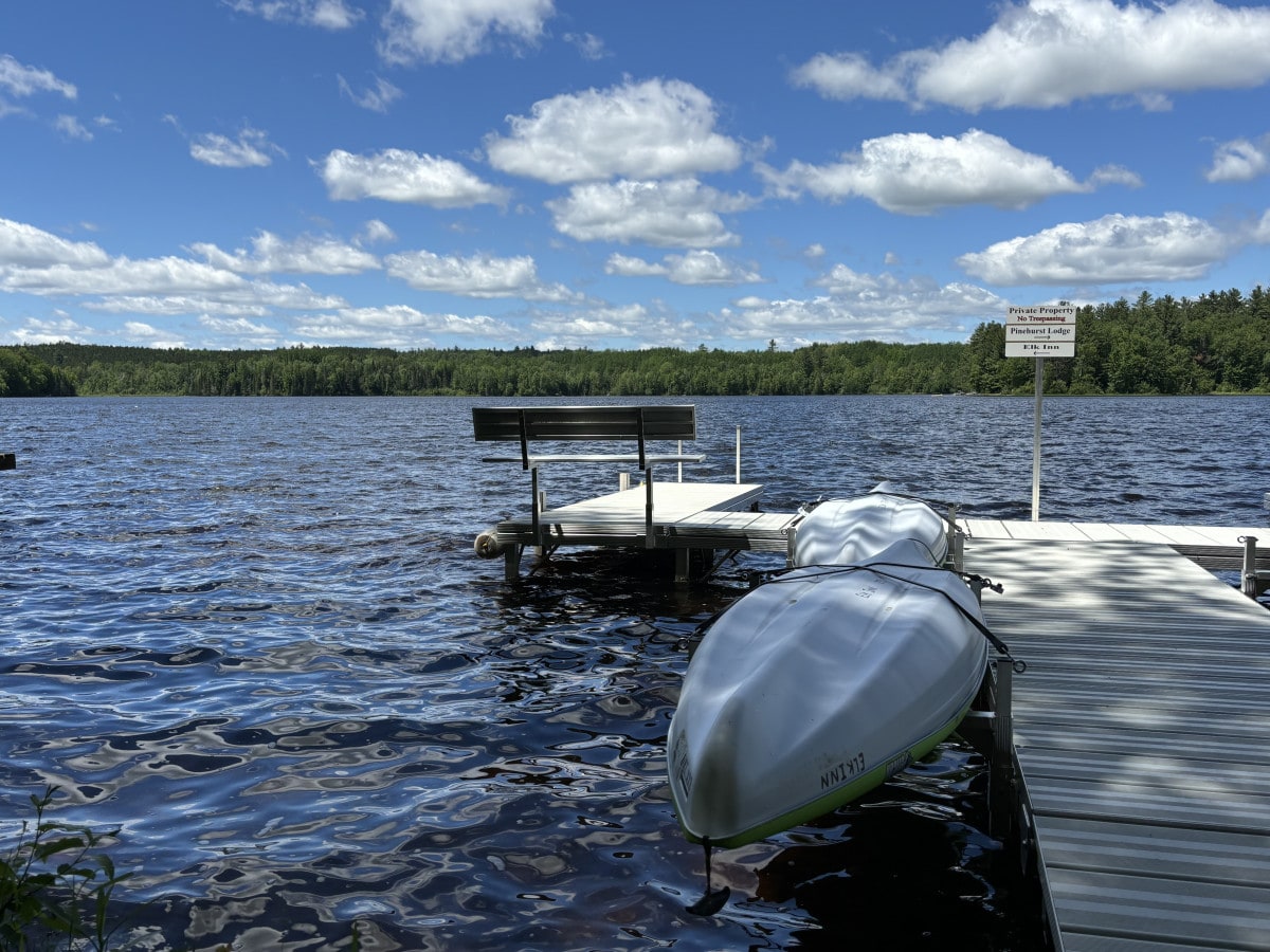 A private dock extends over calm waters, with two kayaks secured at the edge. Lush greenery lines the distant shoreline, and fluffy clouds drift across a blue sky, creating a peaceful setting for outdoor activities.