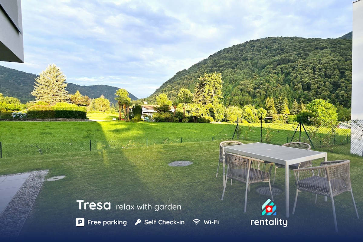 A view of a well-maintained garden area stretches across the image, featuring a spacious lawn surrounded by trees and greenery. Outdoor seating is visible at a dining table, providing a serene space to enjoy the natural surroundings.