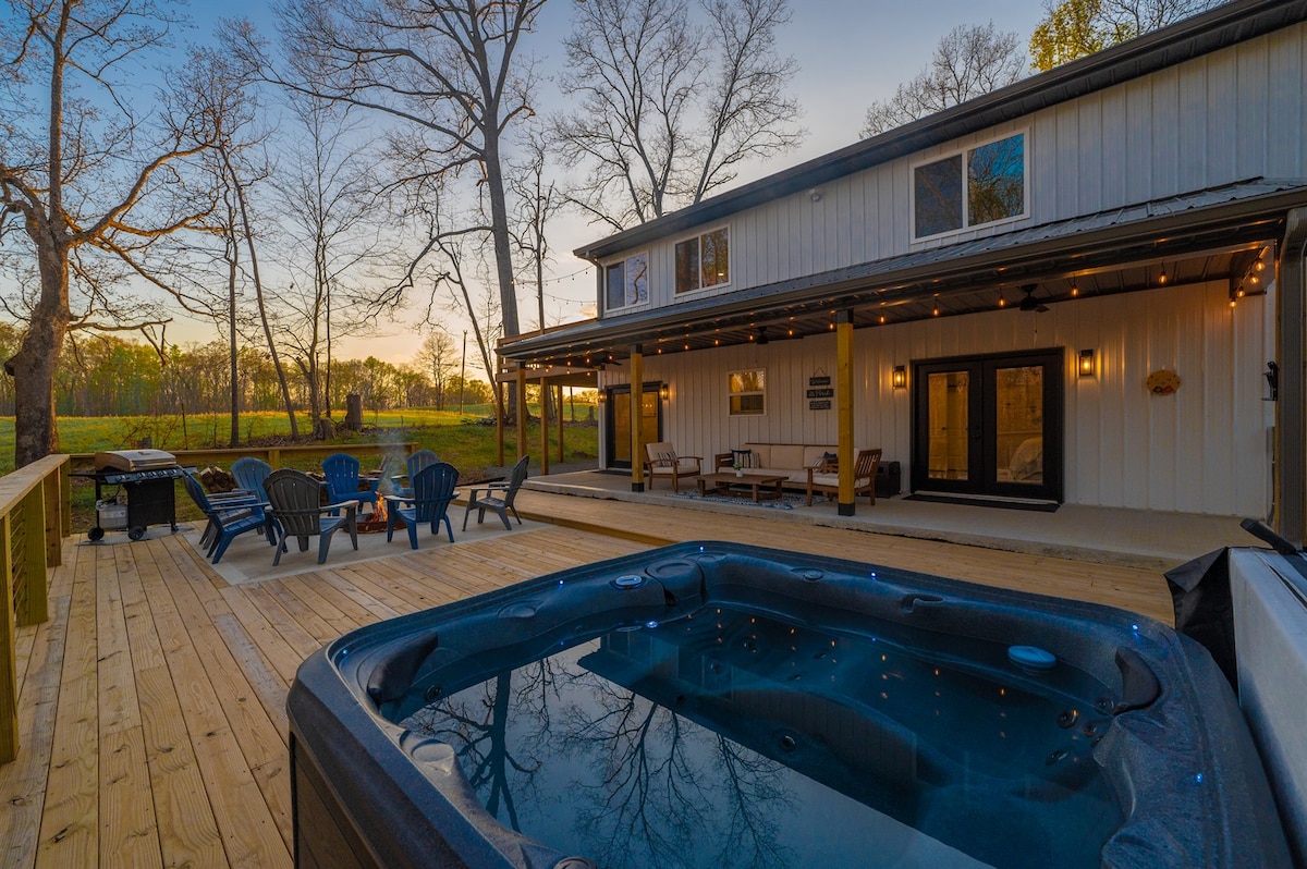 A spacious wooden deck showcases a hot tub with jets, surrounded by outdoor seating, including chairs arranged in a circle. The backdrop features a peaceful landscape of trees and open pasture, with soft evening light enhancing the serene atmosphere.