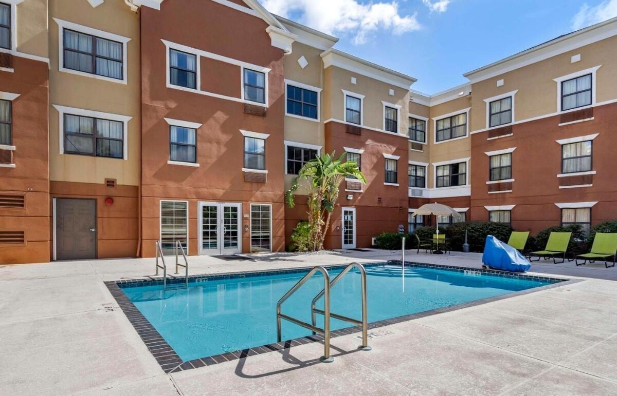 A swimming pool is situated outdoors, surrounded by lounge chairs. The building features a blend of beige and brown siding, with large windows providing natural light. Lush greenery and palm trees accentuate the area, creating a relaxed ambiance.