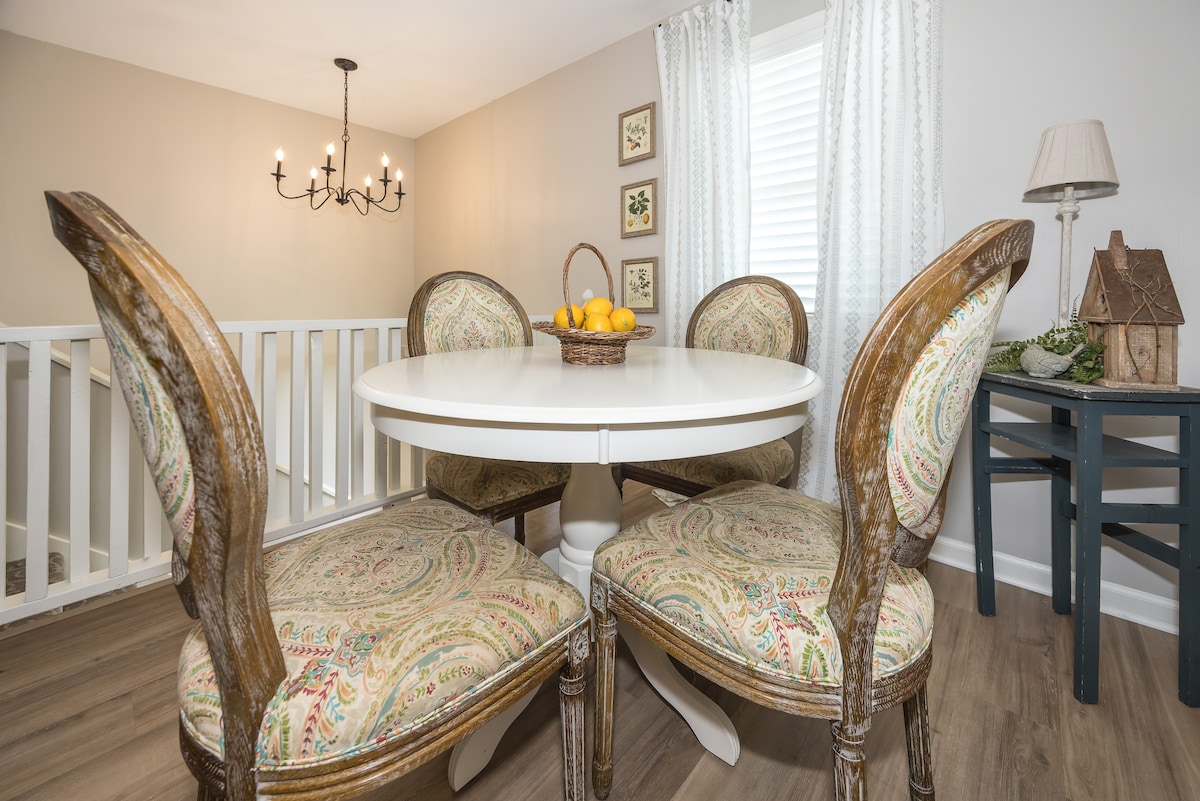 A circular dining table is surrounded by four upholstered chairs featuring intricate patterns. A bowl of lemons sits at the center of the table. The space is illuminated by an elegant chandelier, and natural light filters through sheer curtains at a nearby window.