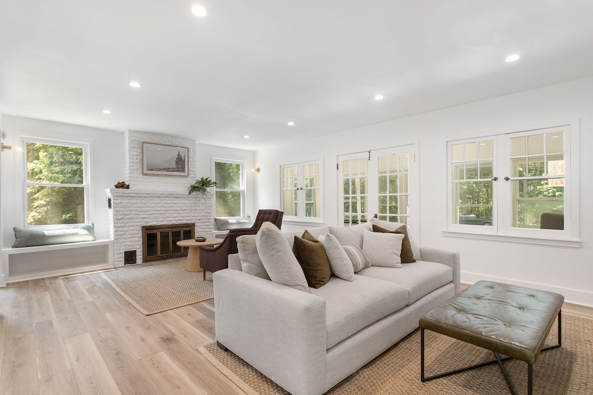 A spacious living room features large windows allowing natural light to fill the space. A comfortable light-colored sofa is complemented by decorative pillows, while a cozy armchair is positioned nearby. A fire place is centered against a white brick wall, adding a touch of warmth.