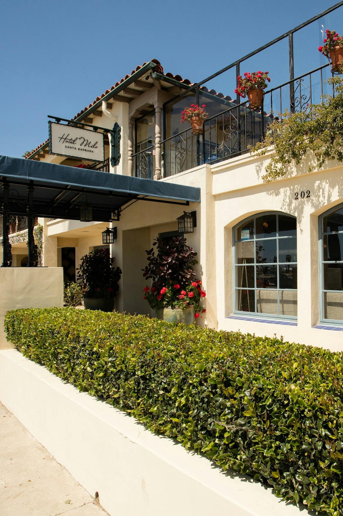 The exterior of the hotel features a Spanish-style architecture with a blue awning and decorative planters filled with flowers. Large windows and a sign displaying the hotel name are prominently displayed, highlighting the inviting entrance surrounded by trimmed hedges.
