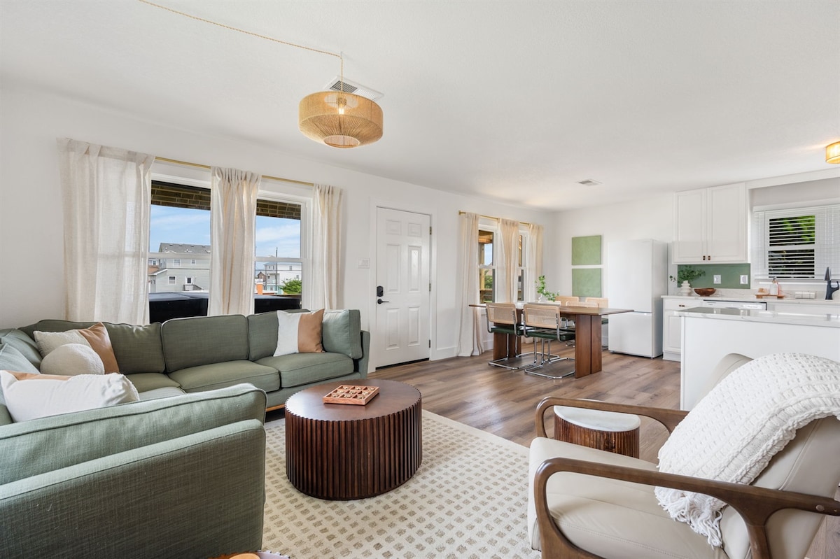 A bright and airy living area is presented, featuring a comfortable green sectional sofa and a round wooden coffee table. Large windows allow for natural light, while a dining area with a small wooden table complements the adjacent kitchen, designed in neutral tones.