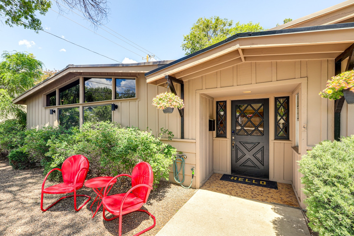The exterior features a welcoming entryway with a dark wood front door and a 'HELLO' mat. Two red chairs are positioned beside the entrance, surrounded by greenery and flower pots. Large windows create a bright and open atmosphere.