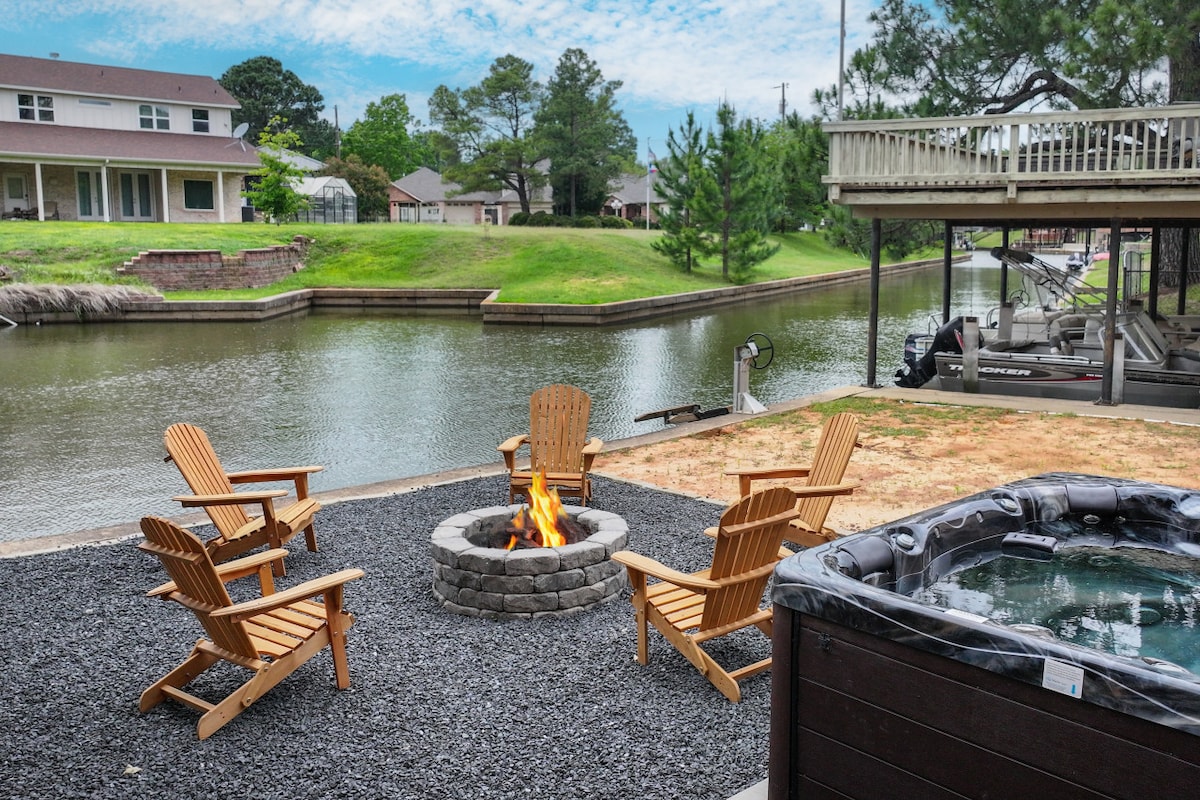 An outdoor gathering area features a stone fire pit surrounded by five wooden chairs, inviting relaxation. Nearby, a hot tub offers a place to unwind while enjoying views of the water. Lush green grass and trees frame the serene canal setting.