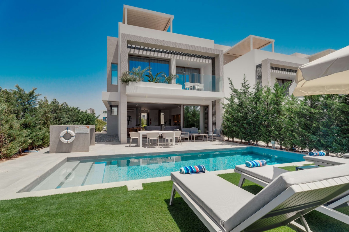 A private pool is set within a spacious yard, surrounded by greenery. Lounge chairs are positioned beside the pool, inviting relaxation. The villa's modern design features large balconies above, enhancing the outdoor living experience under bright blue skies.