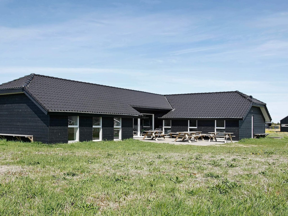 A spacious holiday home is surrounded by greenery, featuring a dark wooden exterior and a large patio area. Multiple picnic tables are arranged outside, providing ample space for outdoor gatherings. Large windows allow natural light to fill the interior, creating a welcoming atmosphere.