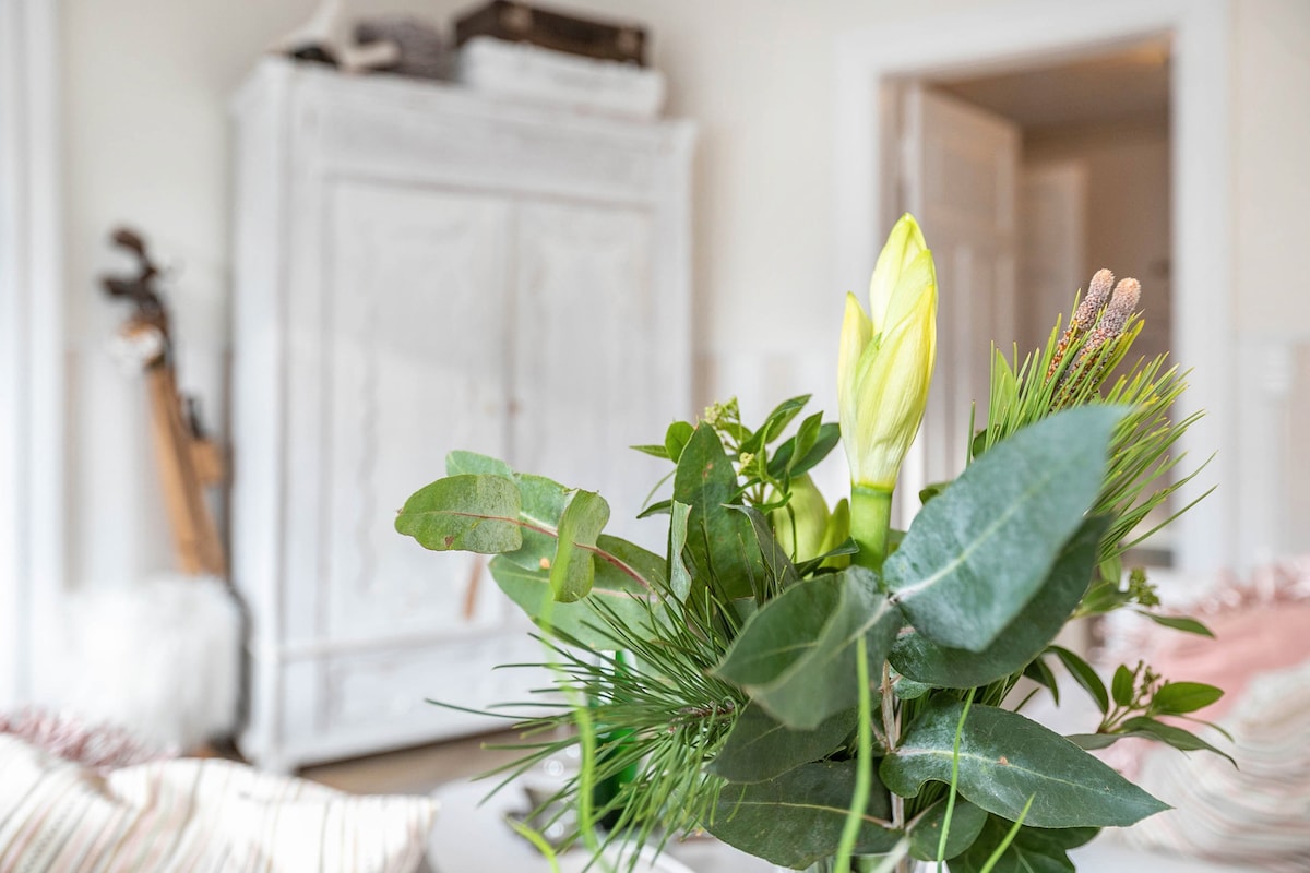 A bouquet featuring green foliage and soft yellow blooms is positioned in the foreground. In the background, a white wooden cabinet stands against a neutral wall, and a patterned bedcover is visible, enhancing the room's comfort.