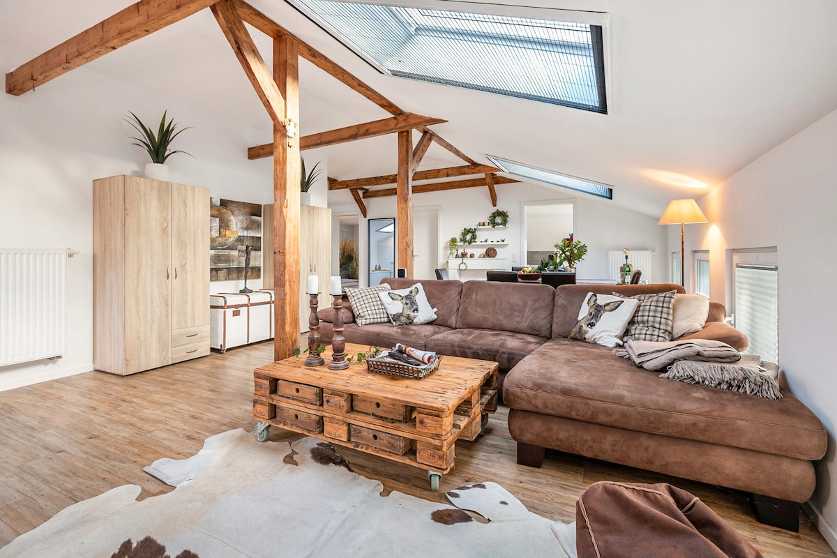 A spacious living area features a large sectional sofa in a neutral tone, complemented by a rustic wooden coffee table. Natural light floods the room through skylights, highlighting wooden beams and stylish decor elements like plants and a soft area rug.