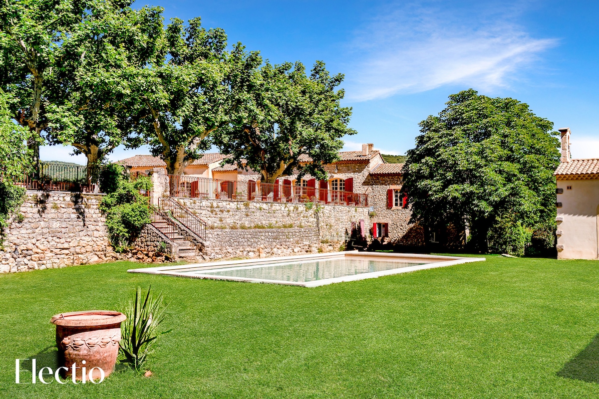 A well-maintained garden surrounds a rectangular swimming pool. Lush greenery and tall trees provide shade, while a stone wall and bright red windows create a charming backdrop. The sunlit landscape invites relaxation, highlighting the tranquility of the estate.
