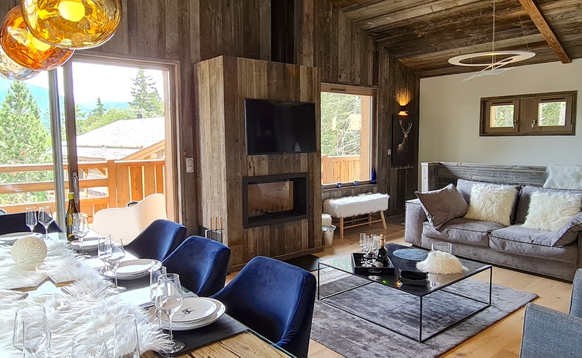 A welcoming living and dining area features a wooden dining table set for a meal, complemented by plush blue chairs. A cozy sofa adorned with soft pillows and a round coffee table is positioned near a modern fireplace, with large windows showcasing views of the surrounding forest.