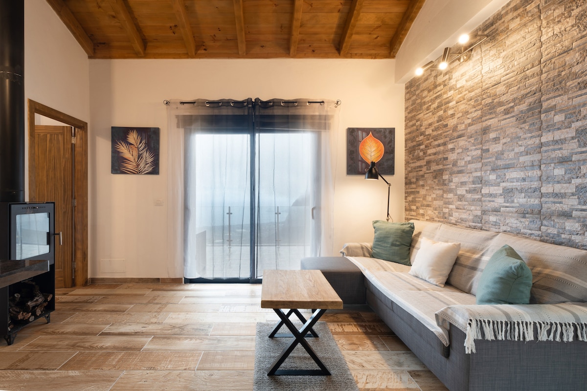 A spacious living area is highlighted by a comfortable sofa with light cushions, positioned near large glass doors that provide access to outdoor views. A wooden coffee table stands in front, complemented by decorative wall art and a cozy fireplace in the corner.