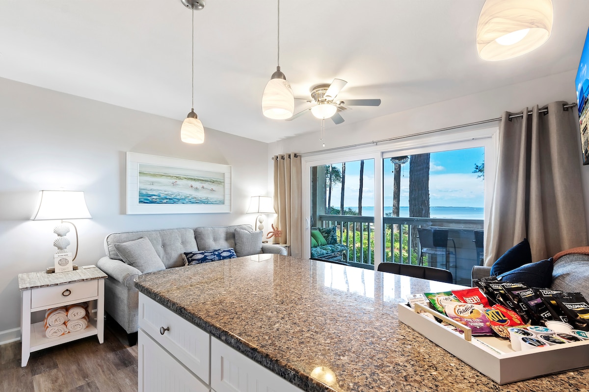 A spacious living area features a light grey sofa complemented by decorative pillows. A large window allows natural light to fill the room, framing views of the outdoors. Pendant lighting hangs above a granite-topped kitchen island, which displays snacks and beverages.