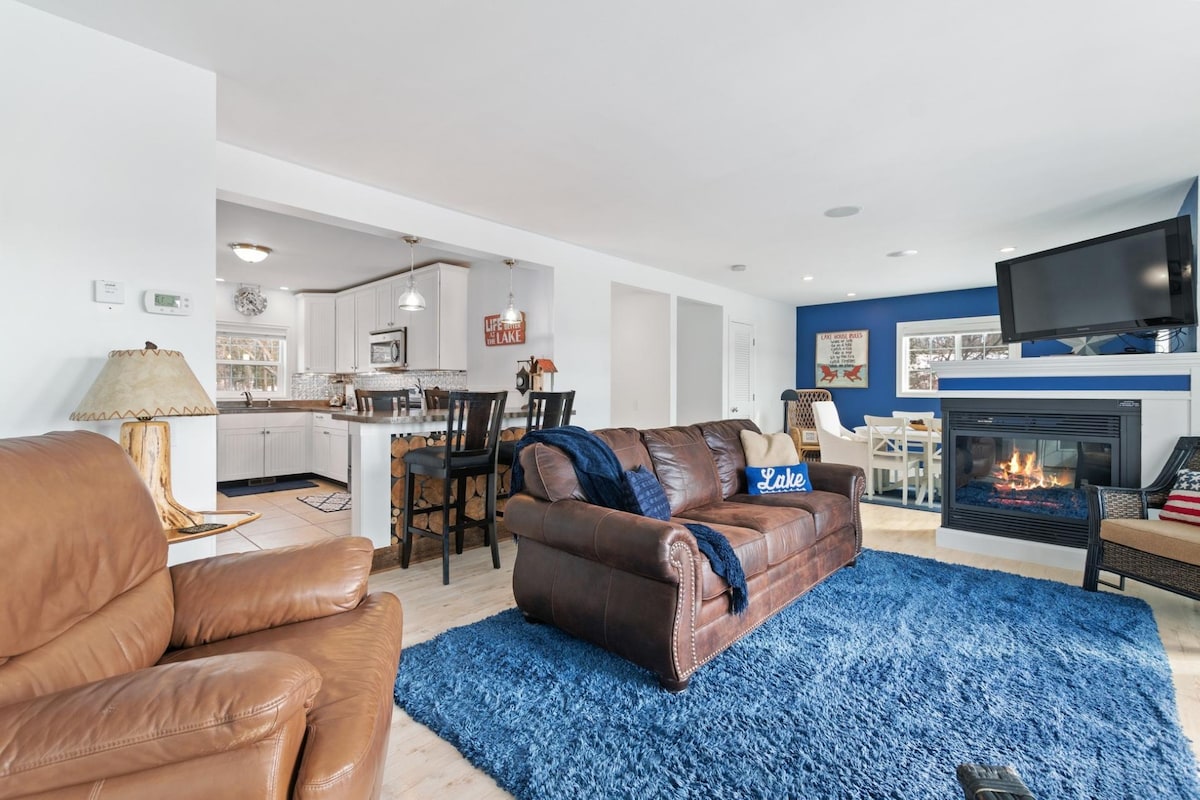 A spacious living area is presented with a large brown leather sofa and two coordinating chairs on a plush blue rug. A cozy fireplace is visible beneath a mounted television, while the open layout provides a clear view of the modern kitchen and dining area.