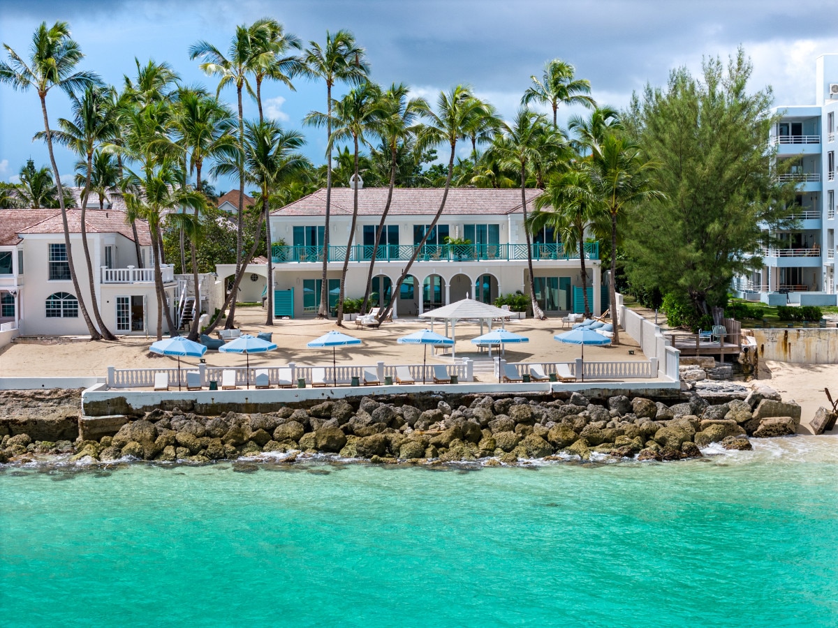 A beachfront property is seen from the water's edge, featuring a two-story structure surrounded by palm trees. Umbrellas and lounge chairs are arranged on the sandy area, with a rocky shore leading to the clear turquoise sea. The building showcases large windows and a welcoming outdoor space.