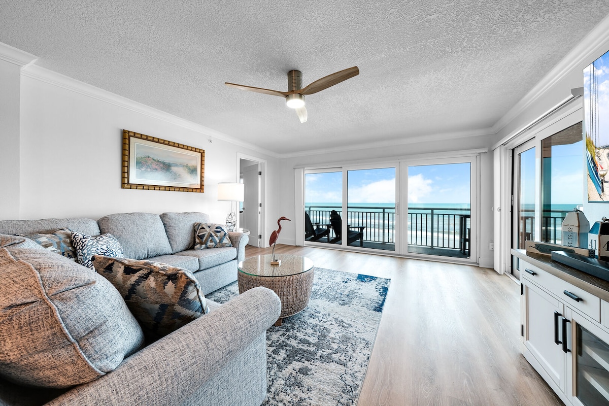 A spacious living area features a comfortable gray sofa and a stylish round table, complemented by a large area rug. Floor-to-ceiling windows provide unobstructed views of the ocean, with a cozy seating area visible on the balcony, enhancing the inviting atmosphere.