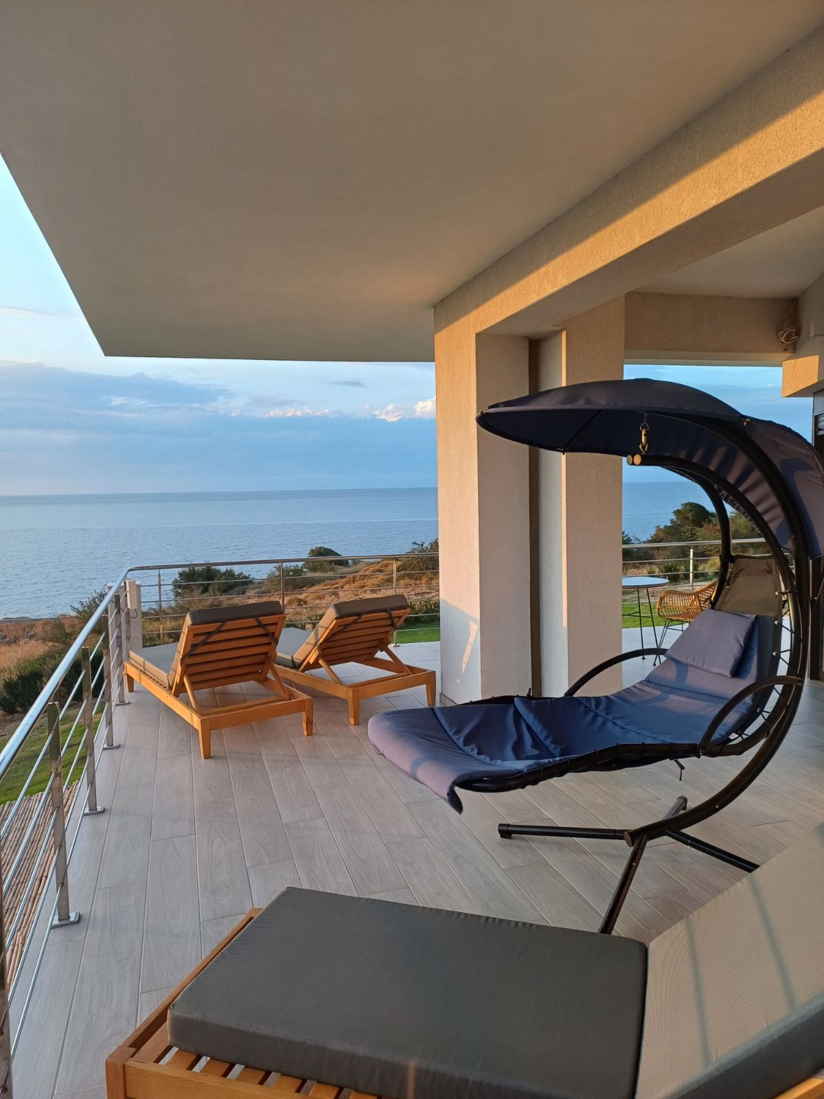 An outdoor terrace features a comfortable hanging chair and two wooden lounge chairs, overlooking a scenic view of the ocean. Soft natural light fills the space, highlighting the smooth decking and the tranquil surroundings.